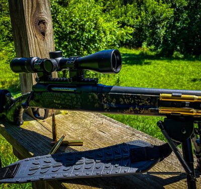 Sniper rifle with mounted scope placed on a wooden platform outdoors, with bullets and a magazine nearby, background featuring green grass and trees.